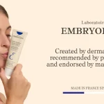 Woman kissing an Embryolisse moisturizer tube in a beige ad backdrop.